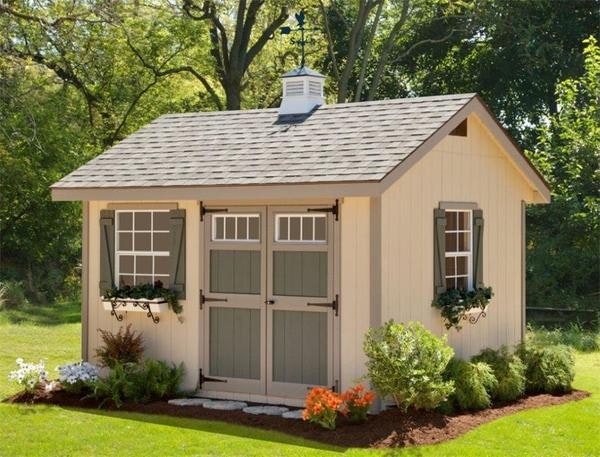 cream colored backyard storage shed with double doors, window boxes, cupola, and landscaped plants
